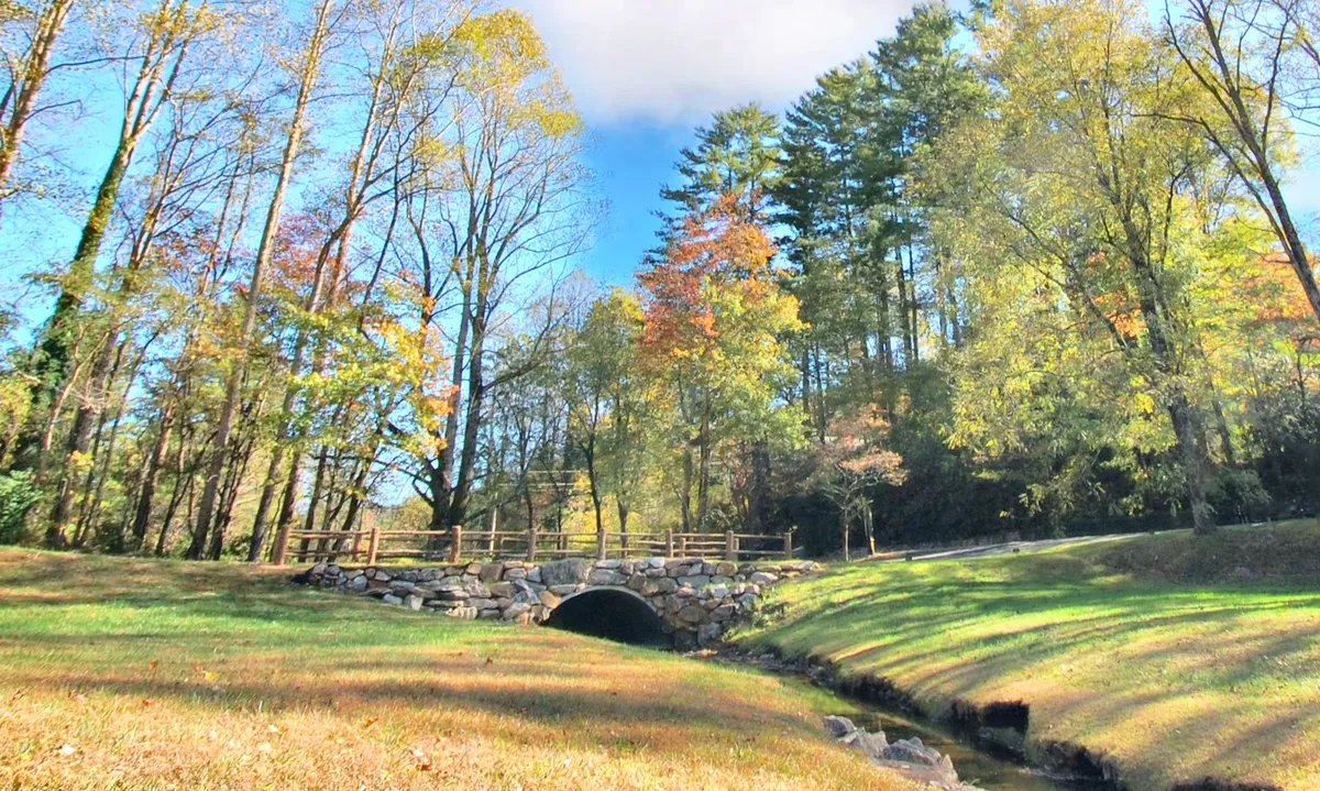 A picturesque stone bridge crosses a quietly meandering creek, surrounded by a tapestry of colorful autumn trees. The scene exudes tranquility and natural beauty, inviting couples to take leisurely walks or enjoy intimate picnics beside the water.
