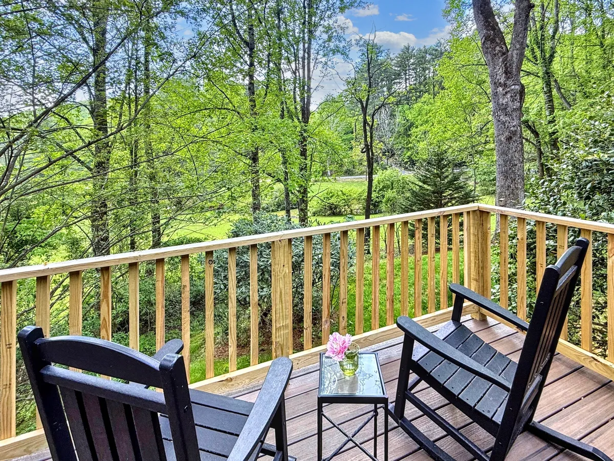 A peaceful wooden deck is outfitted with inviting rocking chairs and a small table adorned with fresh flowers. Lush greenery forms a serene backdrop, suggesting a cozy, private spot to relax, converse, or sip a morning coffee while immersed in nature.