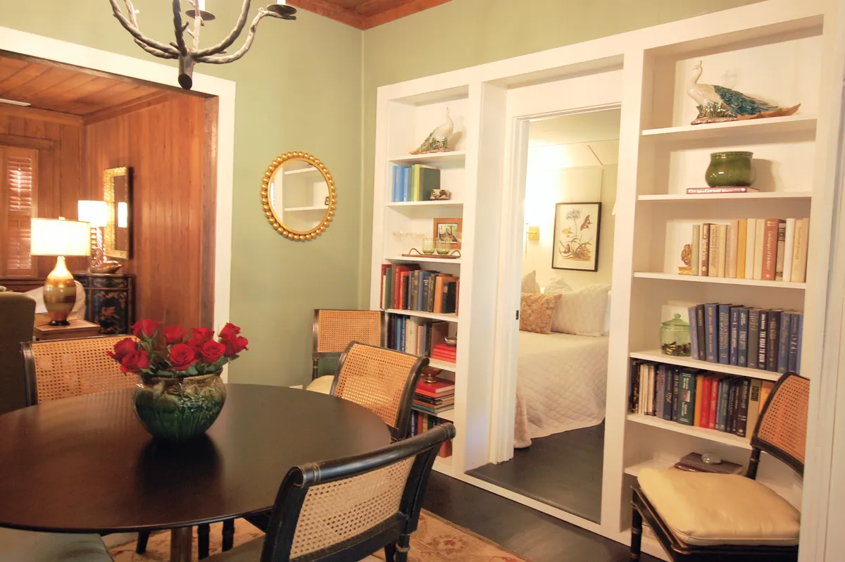 A welcoming dining area with a table set for a meal, surrounded by bookshelves and a mirror that reflects the room’s inviting atmosphere. Sunlight streams in, creating a bright yet cozy setting for socializing and enjoying a candlelit dinner reminiscent of a classic movie scene.