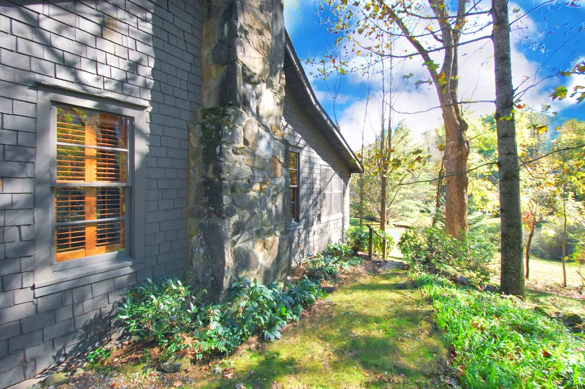 A peaceful garden view featuring a stone wall and windows framed in wood, surrounded by lush greenery and mature trees. The soft natural light and foliage hint at seasonal changes and the serene ambiance of the property’s grounds.