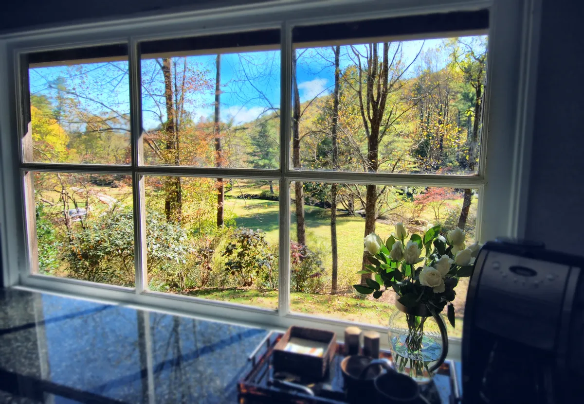 This image looks out a window over a lush garden scene, with a flower arrangement on the windowsill and a coffee machine nearby. Morning light streams in, suggesting a tranquil moment to enjoy the scenery as seasons change. The view beyond the window hints at the property’s connection to nature and the evolving landscape throughout the year.