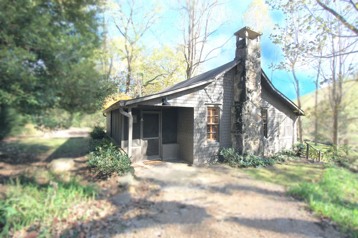 The image captures the exterior of the cottage, highlighting its stone chimney, inviting wooden details, and screened porch. The home is nestled amid lush greenery, creating a welcoming transition between the peacefulness of Horse Cove and easy access to the town's vibrant attractions.