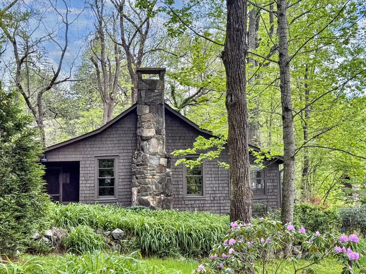 A charming exterior scene showcasing a stone chimney, wooden facade, abundant lush greenery, and flowering plants surrounding the cottage. The environment feels tranquil and inviting, offering easy access to nature just outside the door.