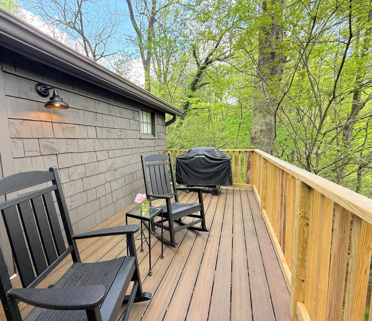 A view of a deck set with two classic rocking chairs and a small table holding a flower arrangement. Surrounded by trees and dappled in soft light, this outdoor space invites guests to relax, enjoy morning coffee, or unwind after a day exploring town.