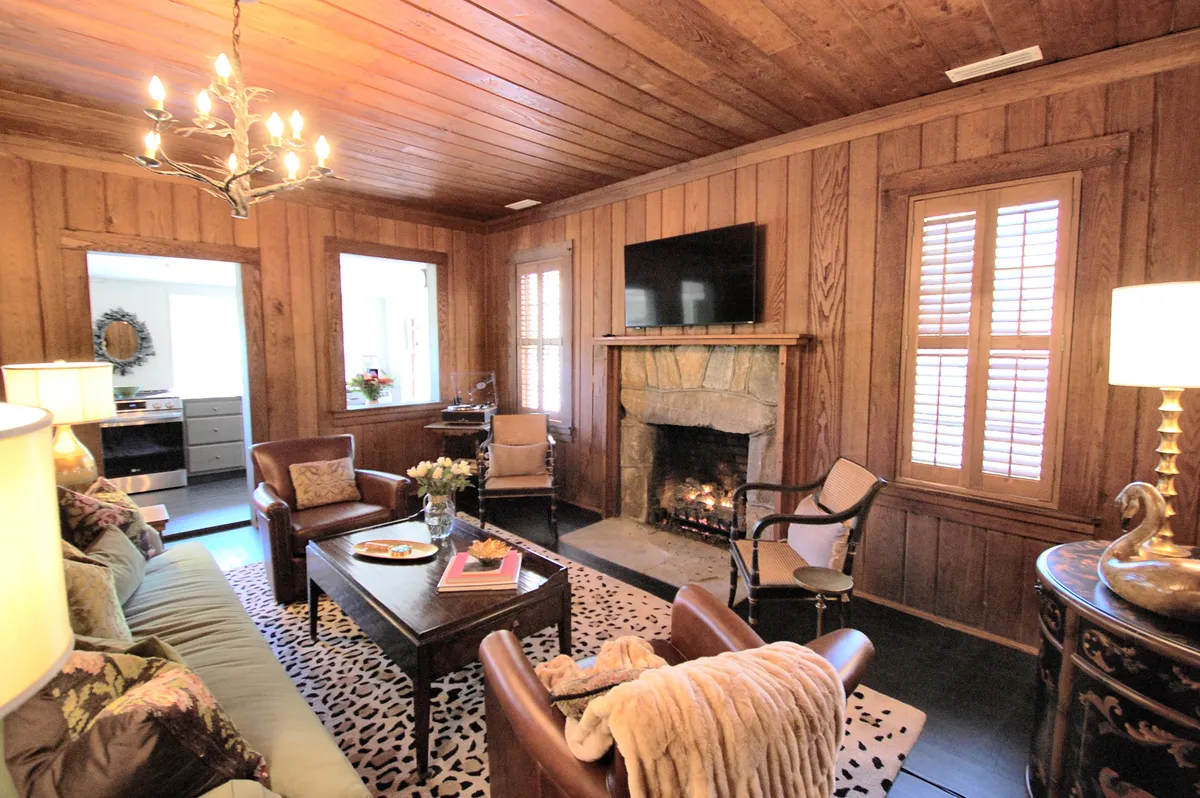 A warmly lit living room featuring a striking stone fireplace as the centerpiece, framed by an elegant chandelier above. Plush, comfortable seating options include a buttery soft sofa and cozy armchairs clustered around a stylish coffee table, with decorative accents on the mantle and side tables enhancing the vintage feel.