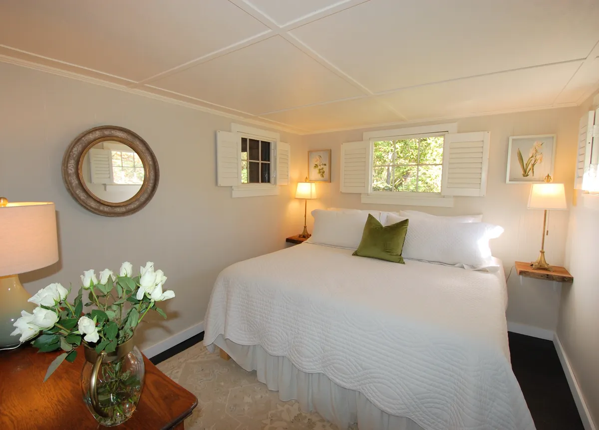 A serene bedroom featuring a plush bed with a white quilt and decorative pillows, flanked by classic nightstands with lamps. A mirror hangs above, and a vase of fresh flowers brings a touch of color. Soft light fills the room, enhancing the feeling of comfort and tranquility.