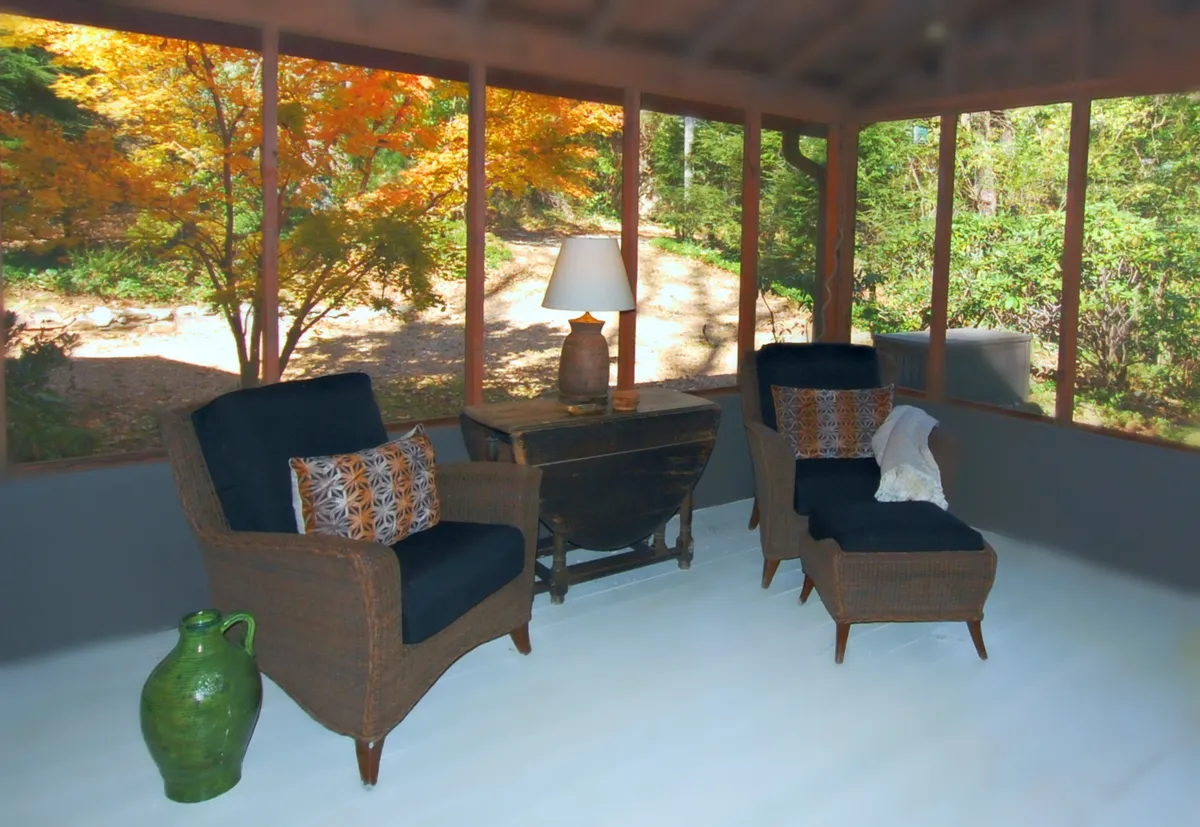 A tranquil sitting area with wicker chairs, decorative cushions, a lamp, and a green vase on a wooden table. The space is softly illuminated, offering views of nature and space for quiet reading or conversation.