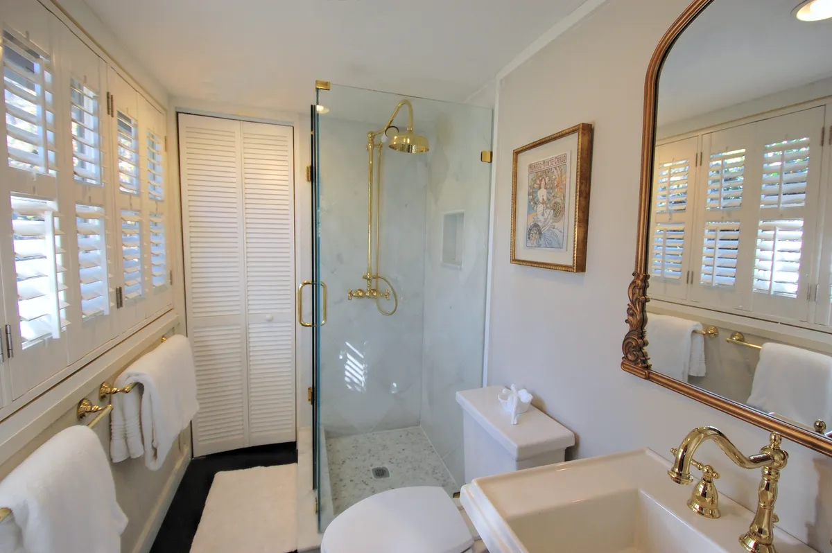A luxurious bathroom featuring a spacious glass shower with gold fixtures, a decorative mirror, and framed artwork. The soft colors and plush finishes evoke the tranquility of a spa, inviting relaxation.
