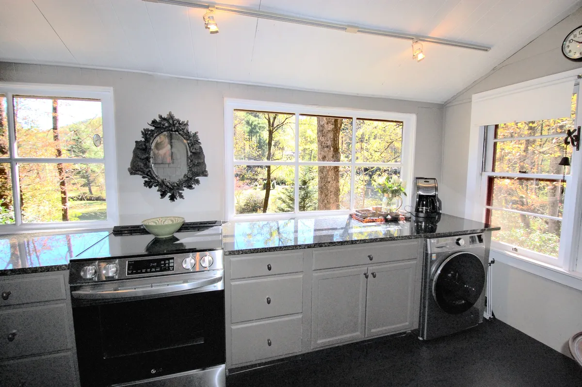A bright, welcoming kitchen featuring a spacious countertop, large windows that flood the space with natural light, a decorative mirror, and a coffee maker. The windows offer charming views of the outdoors, and the kitchen's open design hints at an easy flow into adjacent living spaces.