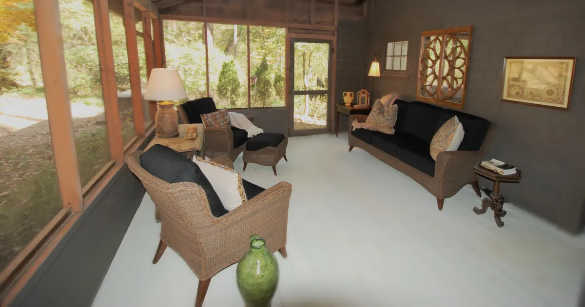 A spacious, light-filled room featuring large windows that let in abundant natural light. Comfortable seating surrounds a decorative wall, with a green ceramic vase adding a touch of nature. Sunlight plays on the walls and floor, creating a tranquil, airy atmosphere.